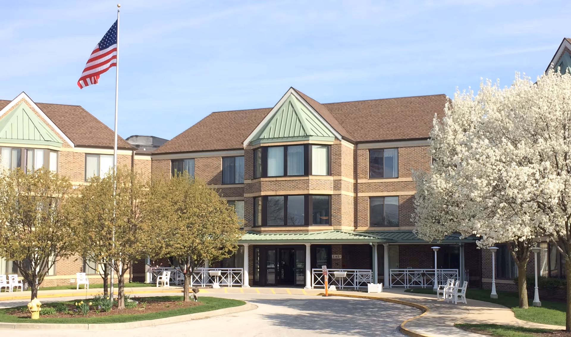 Front exterior view of a three-story brick assisted living facility with green roof accents, an American flag on a flagpole, several trees including one with white blossoms, and a curved driveway leading to the entrance.