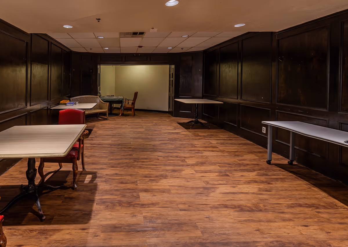 A long, empty interior dining/common area with dark wood paneling, a wooden floor, and several scattered tables and chairs.