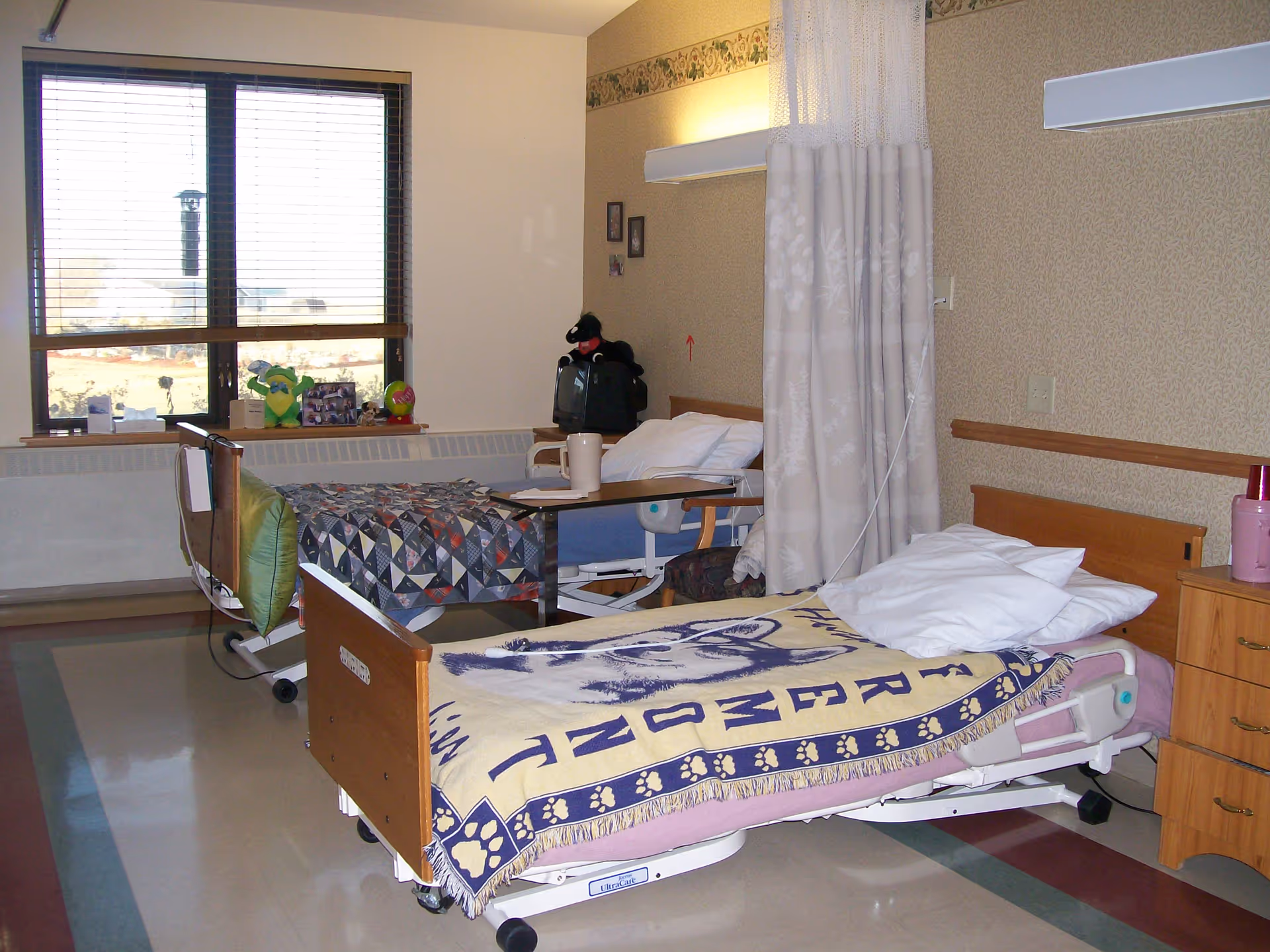 Shared senior living bedroom with two hospital-style beds, a window, and a privacy curtain.