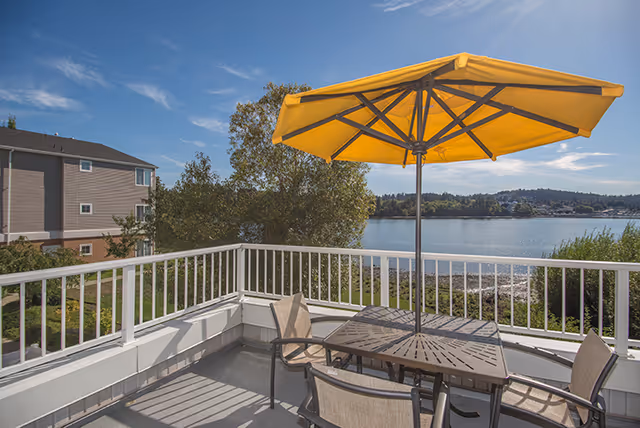 Outdoor patio area with a table and four chairs under a large yellow umbrella, overlooking a body of water with trees and buildings in the background under a clear blue sky.