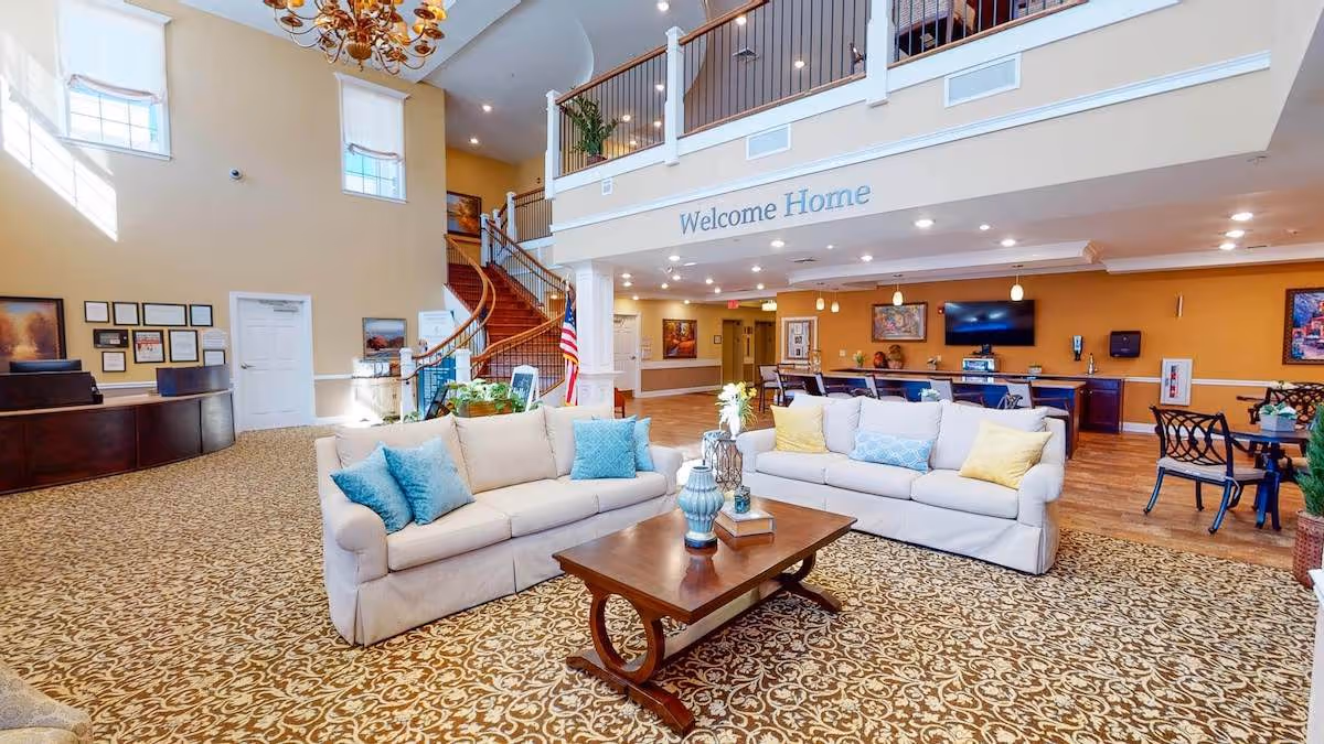 A spacious and well-lit senior living facility common area with two beige sofas adorned with blue and yellow pillows, a wooden coffee table with decorative items, patterned carpet, a curved staircase with wooden handrails, and a reception desk. The wall above the seating area has the words 'Welcome Home'.