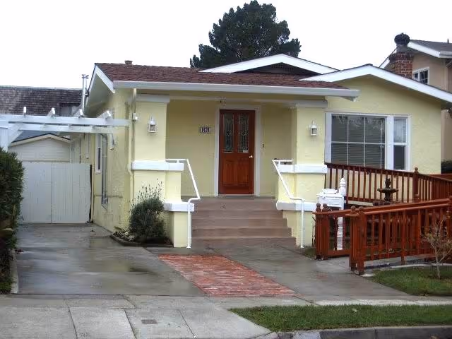 Front exterior view of a single-story yellow house with a brown wooden door, steps leading up to the entrance, white railings, and a wooden ramp on the right side. There is a driveway on the left and a small garden area with shrubs.
