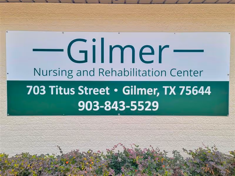 A sign on a beige wall for Gilmer Nursing and Rehabilitation Center, displaying the address 703 Titus Street, Gilmer, TX 75644 and phone number 903-843-5529, with some plants visible at the bottom.