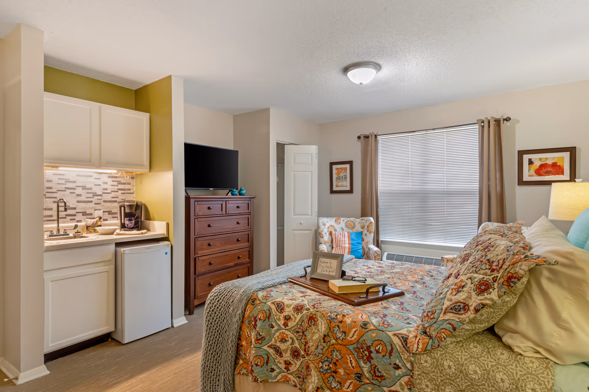 Studio-style bedroom featuring a patterned bed, armchair, dresser with TV, and a kitchenette with a mini-fridge and sink.