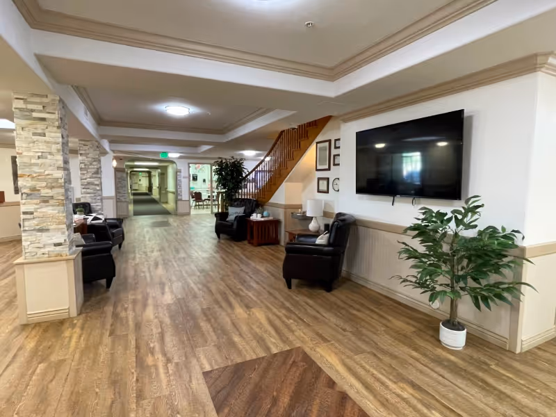 A spacious interior hallway of an assisted living facility with wood flooring, several black armchairs, a large flat-screen TV mounted on the wall, a potted plant, and a staircase leading to an upper floor. The hallway extends into the distance with additional seating and a green exit sign visible.