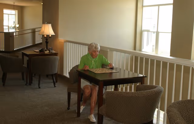 An elderly woman in a green shirt sits at a small table playing a board game in a carpeted lounge area with chairs, a lamp, and large windows.