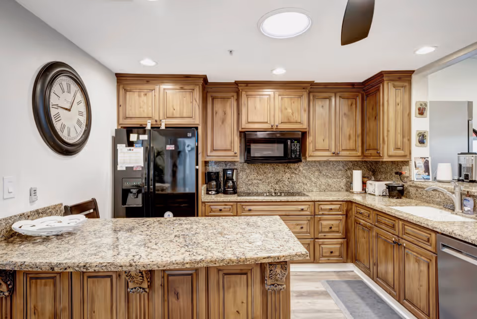 A kitchen with wooden cabinets and granite countertops. The kitchen features a black refrigerator with notes attached, a microwave above a stovetop, two coffee makers, a toaster, a paper towel holder, and a sink with a faucet. A large round wall clock is mounted on the left wall. The floor has light-colored wood or laminate flooring, and there is a kitchen island with ornate wooden detailing.