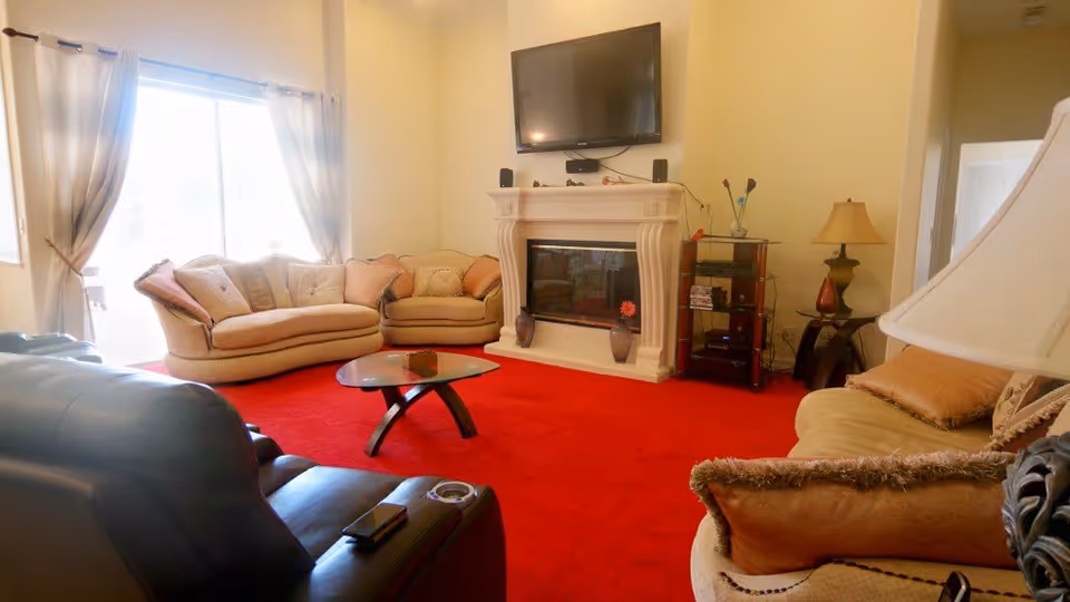 A cozy living room with a bright red carpet, beige sofas with cushions, a glass coffee table, a white fireplace with decorative vases, a flat-screen TV mounted above the fireplace, and a side table with a lamp. Large windows with curtains allow natural light to fill the room.
