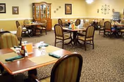 A dining room in a senior living facility with multiple tables and chairs arranged neatly. Each table is set with placemats, napkins, and condiments. The room has carpeted flooring, a wooden china cabinet against the wall, framed artwork, and soft lighting fixtures.