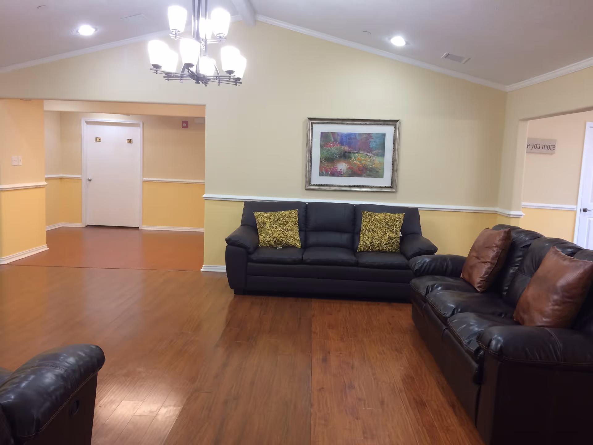 A cozy living room area in an assisted living facility with two black leather sofas, each adorned with decorative pillows. The walls are painted yellow and cream, with a framed floral painting hanging above one sofa. The floor is wooden, and a chandelier with multiple lights hangs from the ceiling. In the background, there are doors labeled 103 and 104.