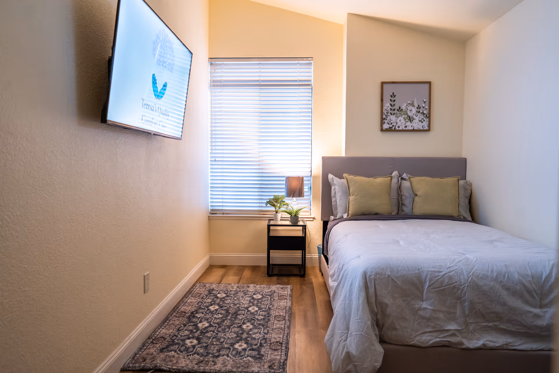 A small, cozy bedroom with a bed featuring a gray headboard, white and green pillows, and a white comforter. There is a window with closed blinds behind a small black nightstand holding a lamp and a plant. A patterned rug lies on the wooden floor, and a flat-screen TV is mounted on the wall opposite the bed. A framed floral artwork hangs on the wall above the bed.