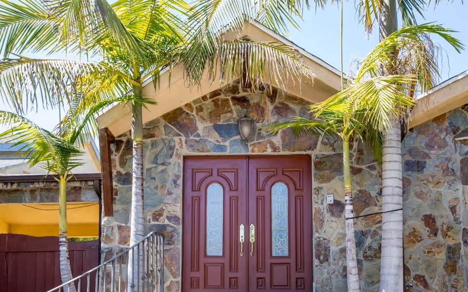Stone facade with maroon double doors and palm trees at the front entrance.