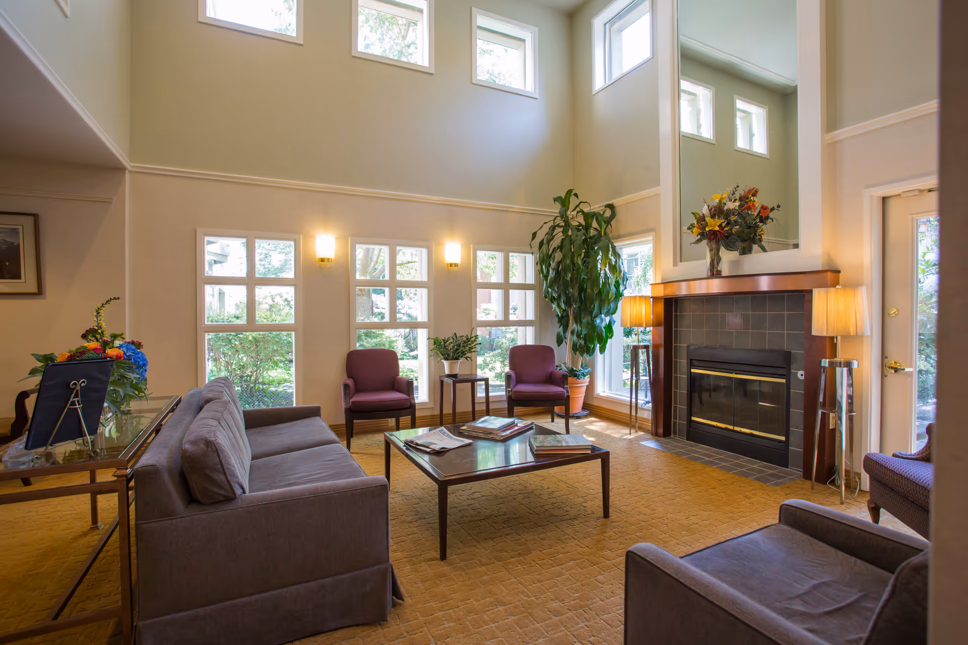 A bright and spacious living room with large windows letting in natural light. The room features a gray sofa, two purple armchairs, a wooden coffee table with magazines, a fireplace with a large mirror above it, two floor lamps, and a tall potted plant. There is also a side table with a colorful flower arrangement.