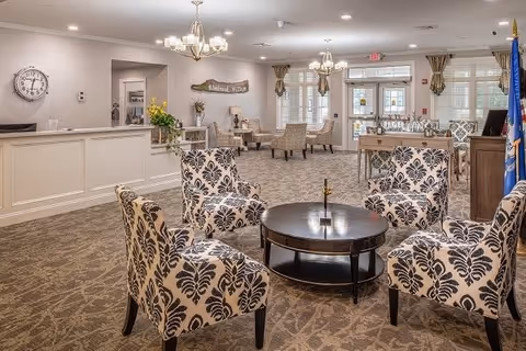A spacious and well-lit common area in a senior living facility featuring four patterned armchairs arranged around a round wooden coffee table. The room has beige walls, carpeted floors, and multiple chandeliers hanging from the ceiling. In the background, there is a reception desk, additional seating with armchairs, large windows with curtains, and a door leading outside. Decorative elements include a wall clock, plants, and a sign that reads 'Elmbrook Village'.