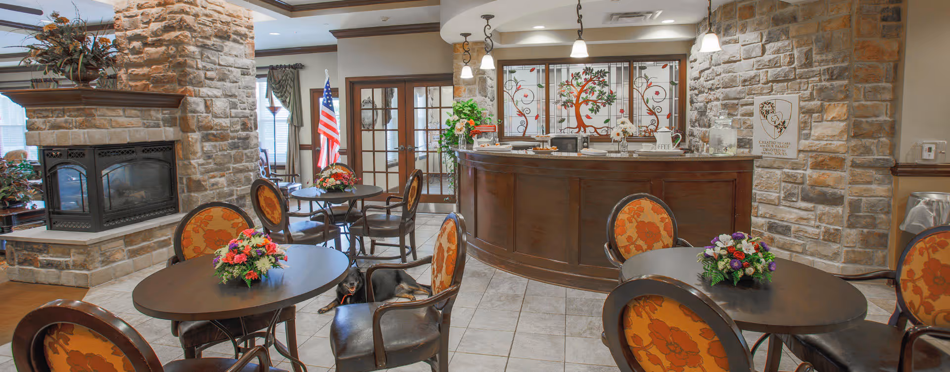 A cozy common area with round tables and chairs featuring orange floral upholstery. Each table has a colorful flower arrangement. There is a stone fireplace on the left side and a curved wooden reception desk with decorative glass panels behind it. An American flag is visible near the windows with curtains, and the floor is tiled.