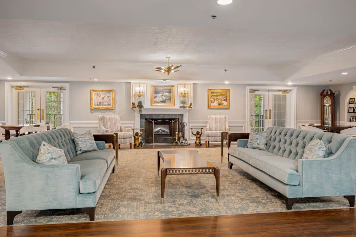 A spacious and elegant living room in Cumberland Hills Assisted Living featuring two light blue tufted sofas facing each other with a wooden coffee table in between. The room has a fireplace with a decorative screen, flanked by two armchairs and wall sconces. There are framed paintings above the fireplace and double glass doors on either side leading outside. The floor is a combination of wood and a large patterned area rug.
