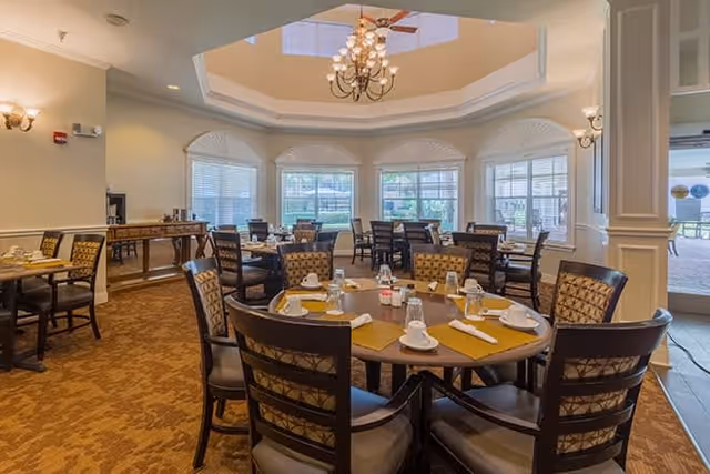 A well-lit dining room with multiple round tables set with cups, glasses, napkins, and condiments. The room features large windows with white blinds, a decorative chandelier hanging from a recessed ceiling, and upholstered chairs around the tables. There is a sideboard against the wall and an open doorway leading to an outdoor patio area.