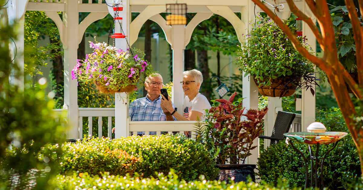 An elderly man and woman sitting together in a white wooden gazebo surrounded by lush greenery and hanging flower baskets. The woman is holding a smartphone and smiling, while the man looks ahead. The scene is bright and peaceful, with various plants and trees around the gazebo.