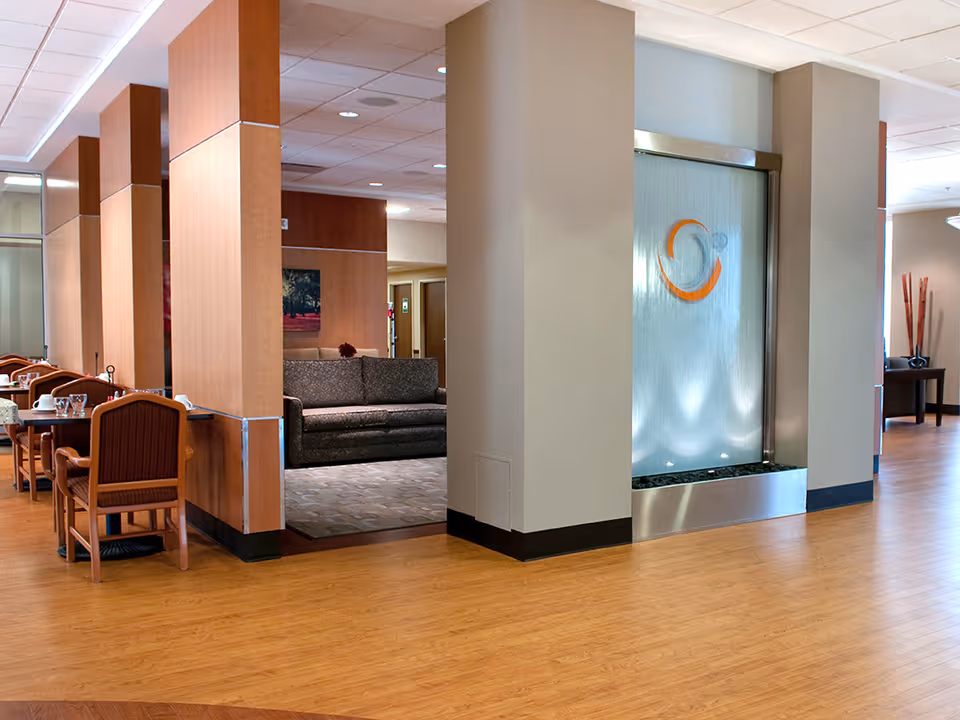 Interior view of a senior living facility showing a seating area with a gray couch, wooden dining tables with chairs, and a decorative water feature with an orange and gray circular design on a frosted glass panel. The floor is wood, and the walls are light-colored with wooden accents.