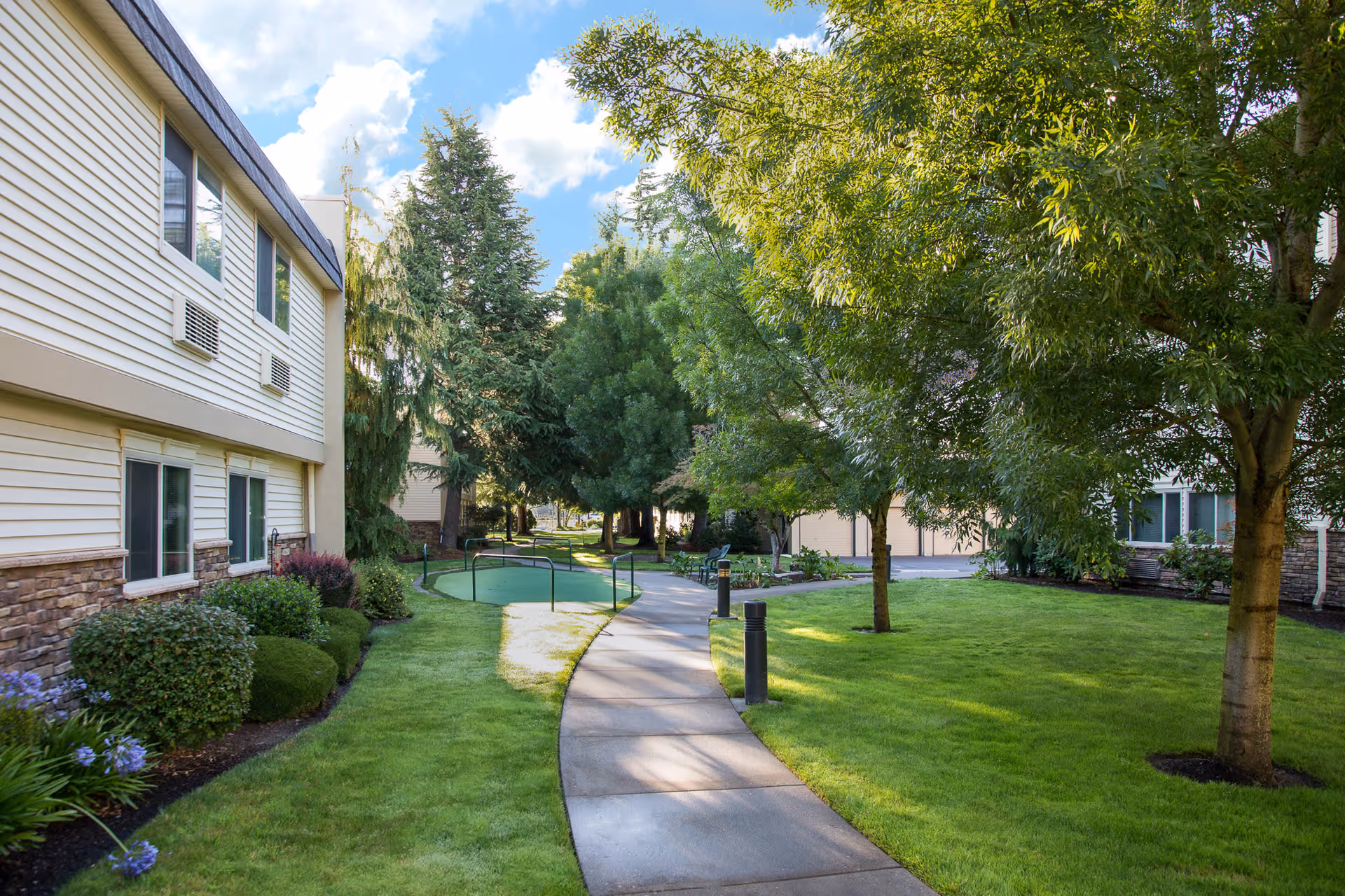 A paved walkway winds through a well-maintained green lawn with trees and shrubs on either side, adjacent to a two-story building with beige siding and stone accents under a partly cloudy blue sky.
