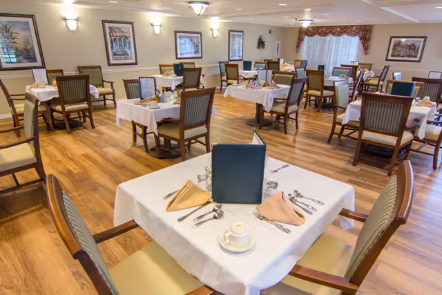 A spacious dining room with multiple tables covered in white tablecloths, each set with folded napkins, silverware, cups, and menus. The room has wooden flooring, framed artwork on the walls, and large windows with curtains allowing natural light to enter.