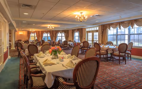 A spacious dining room with multiple round and rectangular tables covered with white tablecloths, set with napkins, plates, and glassware. The room features elegant wooden chairs with striped upholstery, large windows with gold curtains allowing natural light, chandeliers hanging from a white ceiling, and a patterned carpet floor.