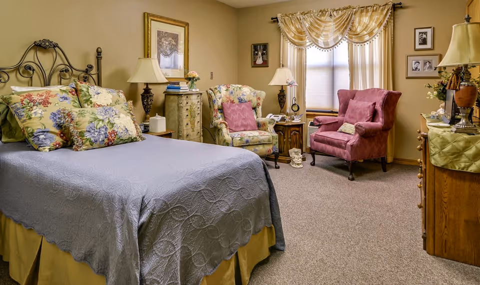 A cozy bedroom in a senior living facility featuring a bed with floral pillows and a gray quilt, two upholstered armchairs (one floral and one pink), a wooden dresser with a lamp and flowers, framed pictures on the walls, and a window with sheer curtains and valance.
