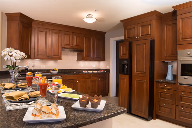 A spacious kitchen with dark wood cabinets, granite countertops and an island set with plates of sandwiches, pastries and drinks.