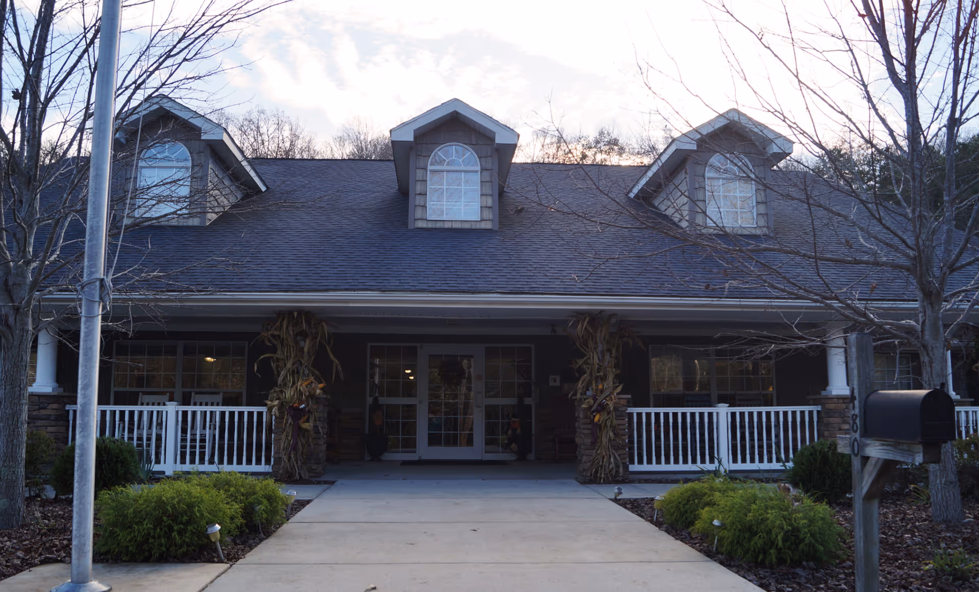 Front exterior view of a residential-style building with a covered porch, white railings, and three dormer windows on the roof. The entrance is decorated with dried corn stalks on either side of the door. There are leafless trees and green shrubs in the landscaped area near the walkway leading to the entrance.