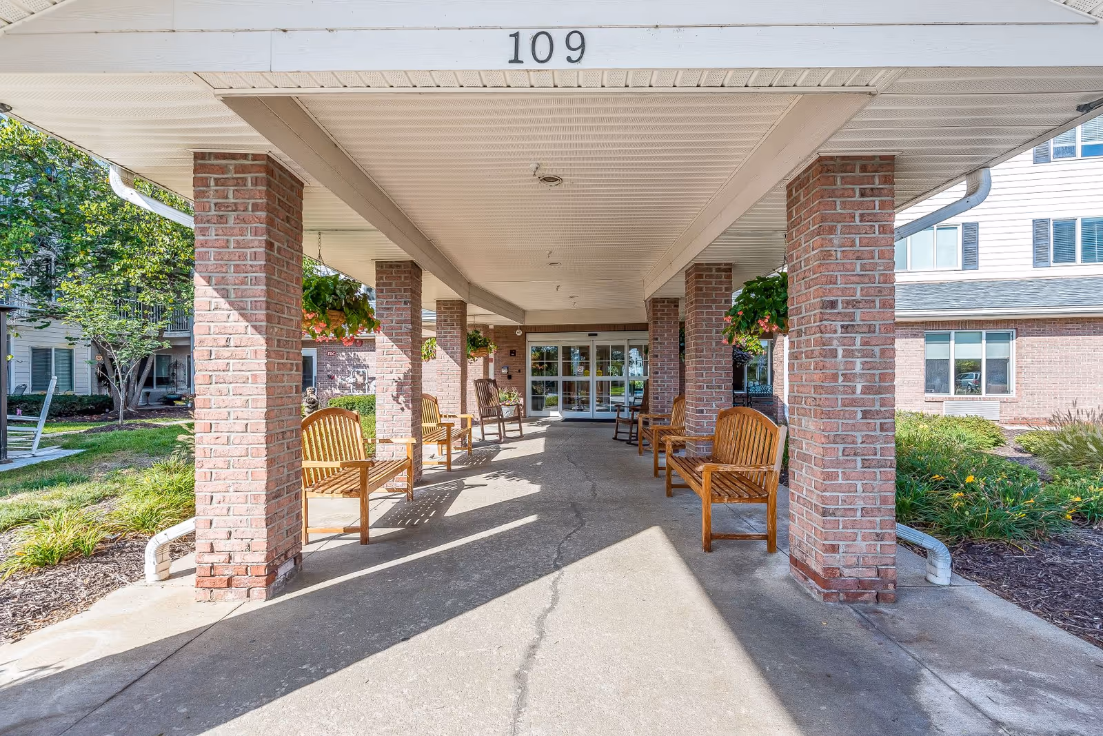 Covered entrance area of a senior living facility with brick pillars and wooden benches on both sides. Hanging flower baskets are attached to the pillars. The building entrance with glass doors is visible at the end of the walkway. The number 109 is displayed on the ceiling above the entrance.