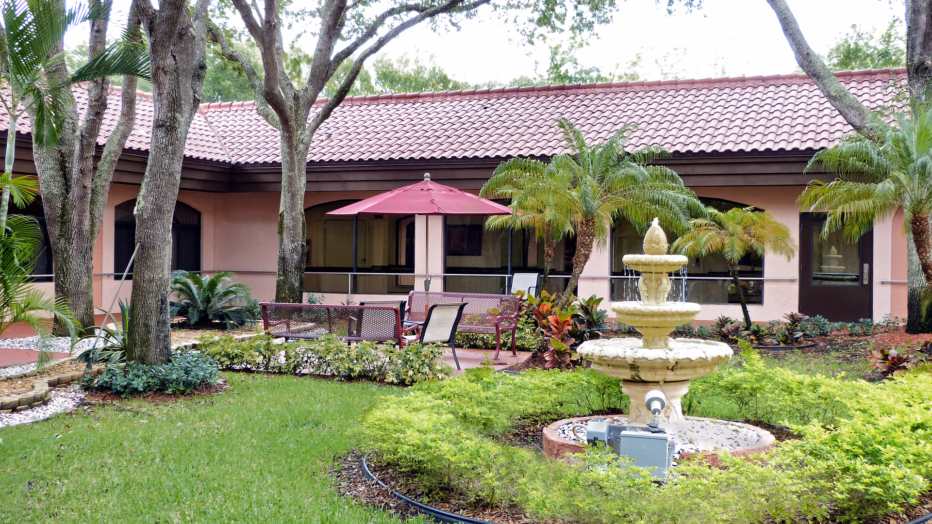 Outdoor courtyard area with green grass, trees, and a three-tiered water fountain surrounded by plants. There is a seating area with chairs and a table under a red umbrella. The building with a tiled roof and windows is visible in the background.