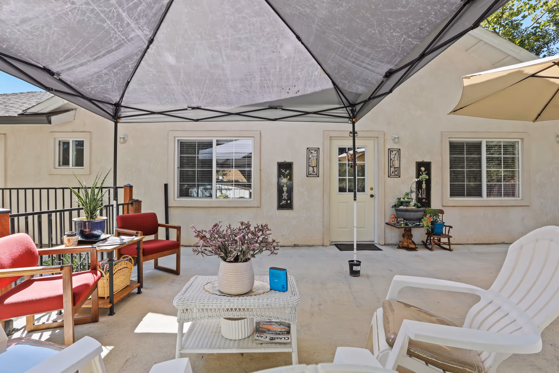 Outdoor patio area at Citrus Pines Senior Living with a canopy overhead, red cushioned chairs, a white wicker coffee table with a potted plant, and a white plastic chair. The patio is adjacent to a beige building with two windows and a door, decorated with wall art and additional potted plants.