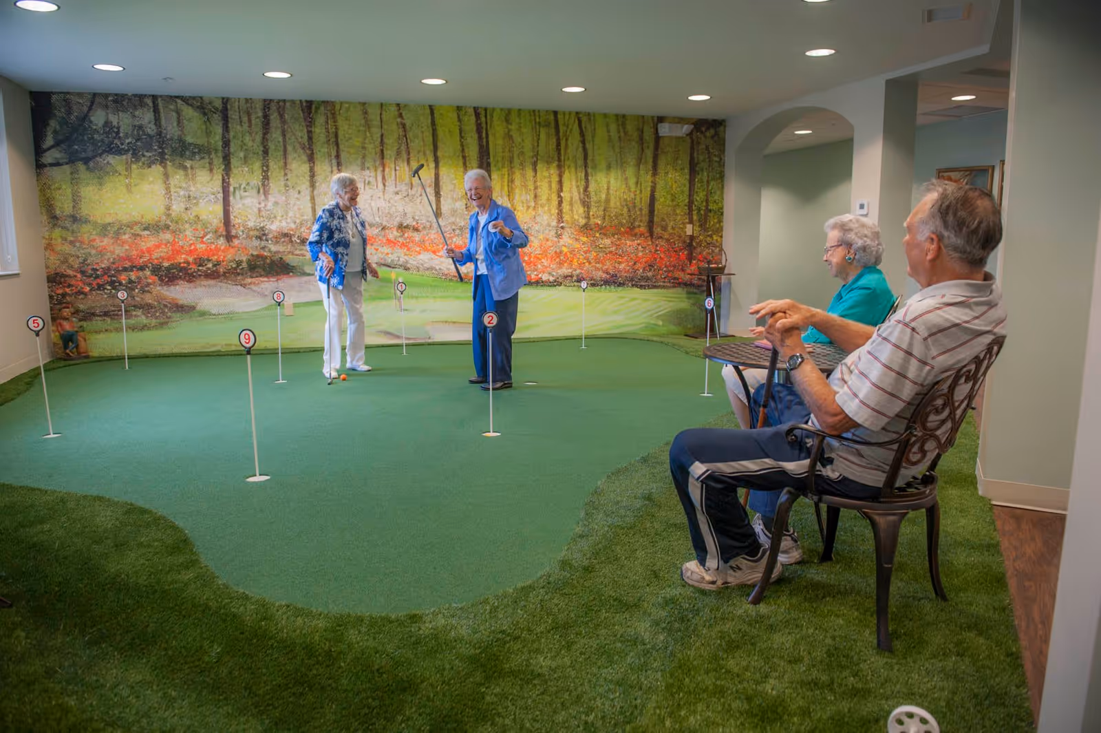Two elderly women playing indoor mini golf on a putting green with numbered holes, while an elderly man and woman sit nearby on metal chairs watching and enjoying the activity. The room has a large mural of a forest scene on the back wall.