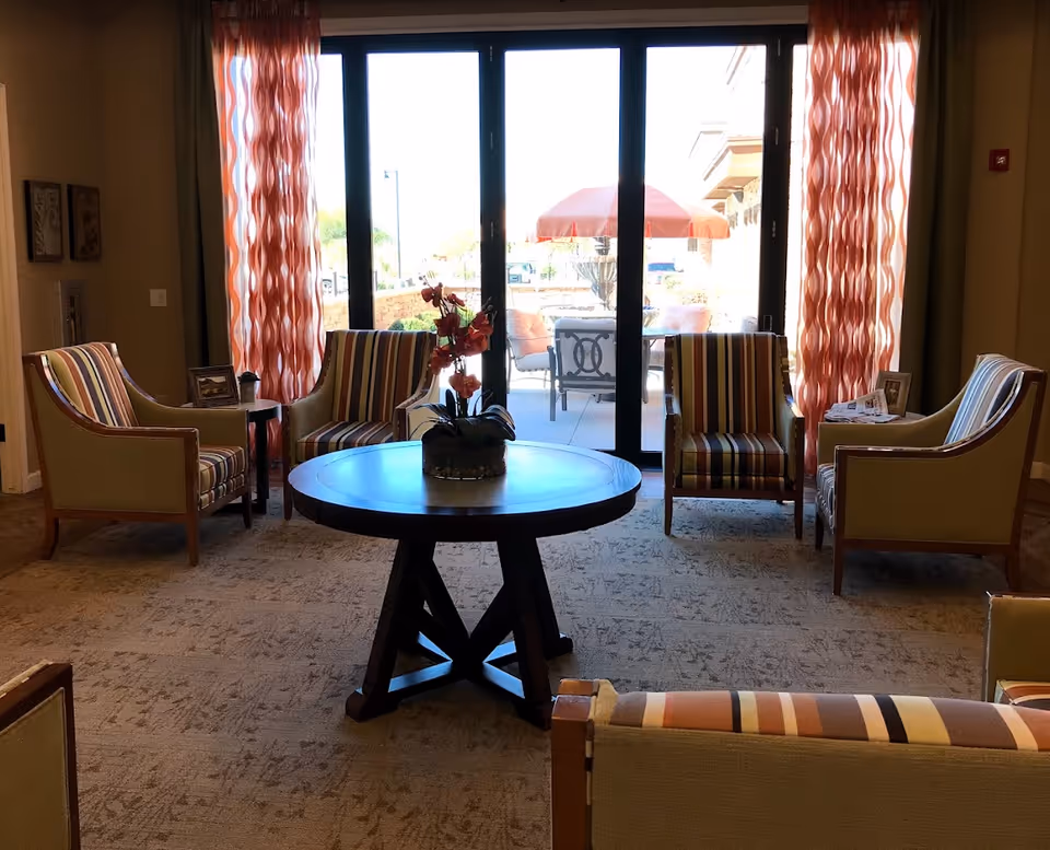 Sunlit seating area with a round central table, striped armchairs, and sliding glass doors opening to a patio.