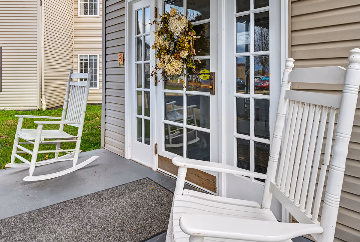 Two white wooden rocking chairs on a porch outside a building entrance with glass-paneled double doors decorated with a floral wreath. The porch has a gray floor and a dark doormat, with beige siding on the building and a grassy area visible in the background.