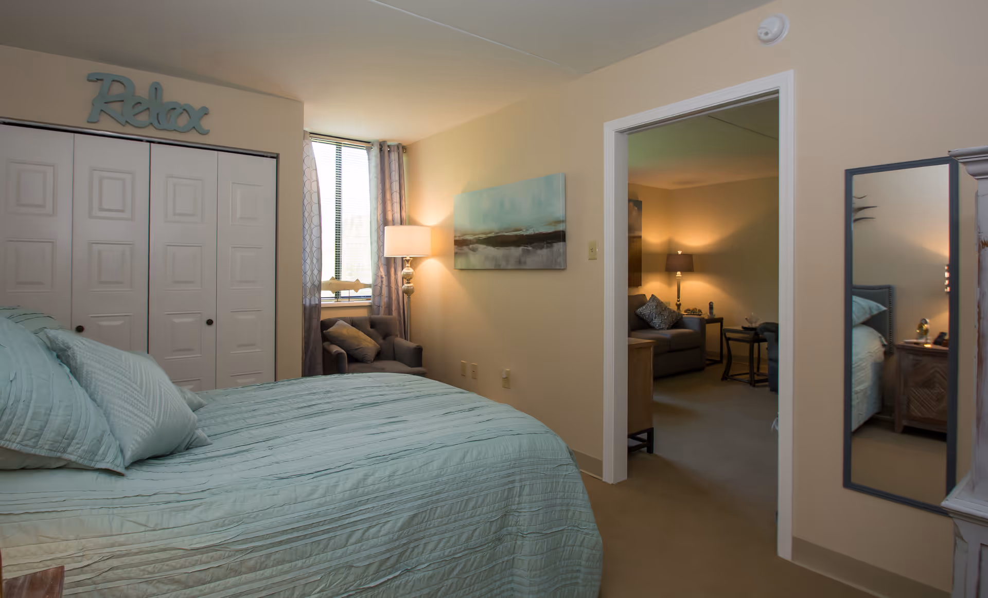 A cozy bedroom with a neatly made bed covered in light blue bedding. There is a window with curtains and a small armchair next to it. Above the closet doors, a decorative sign reads 'Relax'. The room opens into a softly lit living area with a sofa, side table, and lamp. A full-length mirror is mounted on the wall near the doorway.
