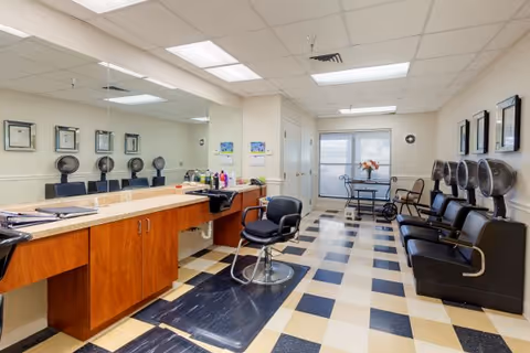 Interior view of a hair salon area in an assisted living facility with a large mirror, a styling chair, hair washing stations with black chairs and sinks, and a small table with chairs near a window at the far end.