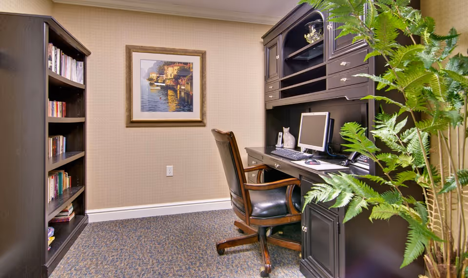 A small office area with a dark wooden desk and matching chair on wheels. The desk has a computer monitor, keyboard, mouse, and a few decorative items. To the left, there is a tall bookshelf filled with books. A framed painting of a waterfront scene hangs on the beige wall. A green leafy plant is visible on the right side of the image.