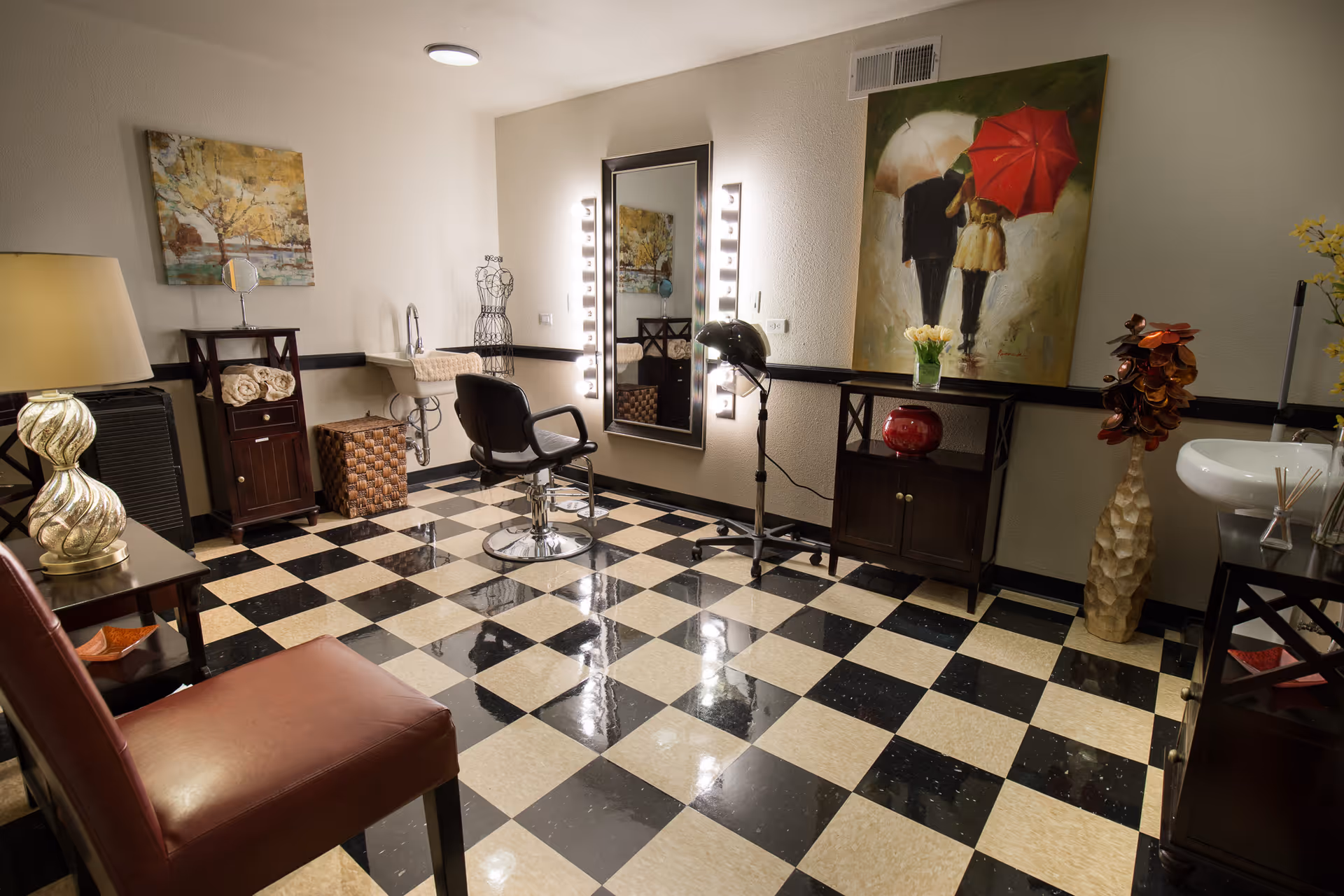 Interior view of a well-lit salon or beauty room with a black and white checkered floor. The room features a salon chair in front of a large mirror surrounded by lights, a hair dryer on a stand, a small sink, decorative paintings on the walls, and various pieces of furniture including cabinets and chairs.