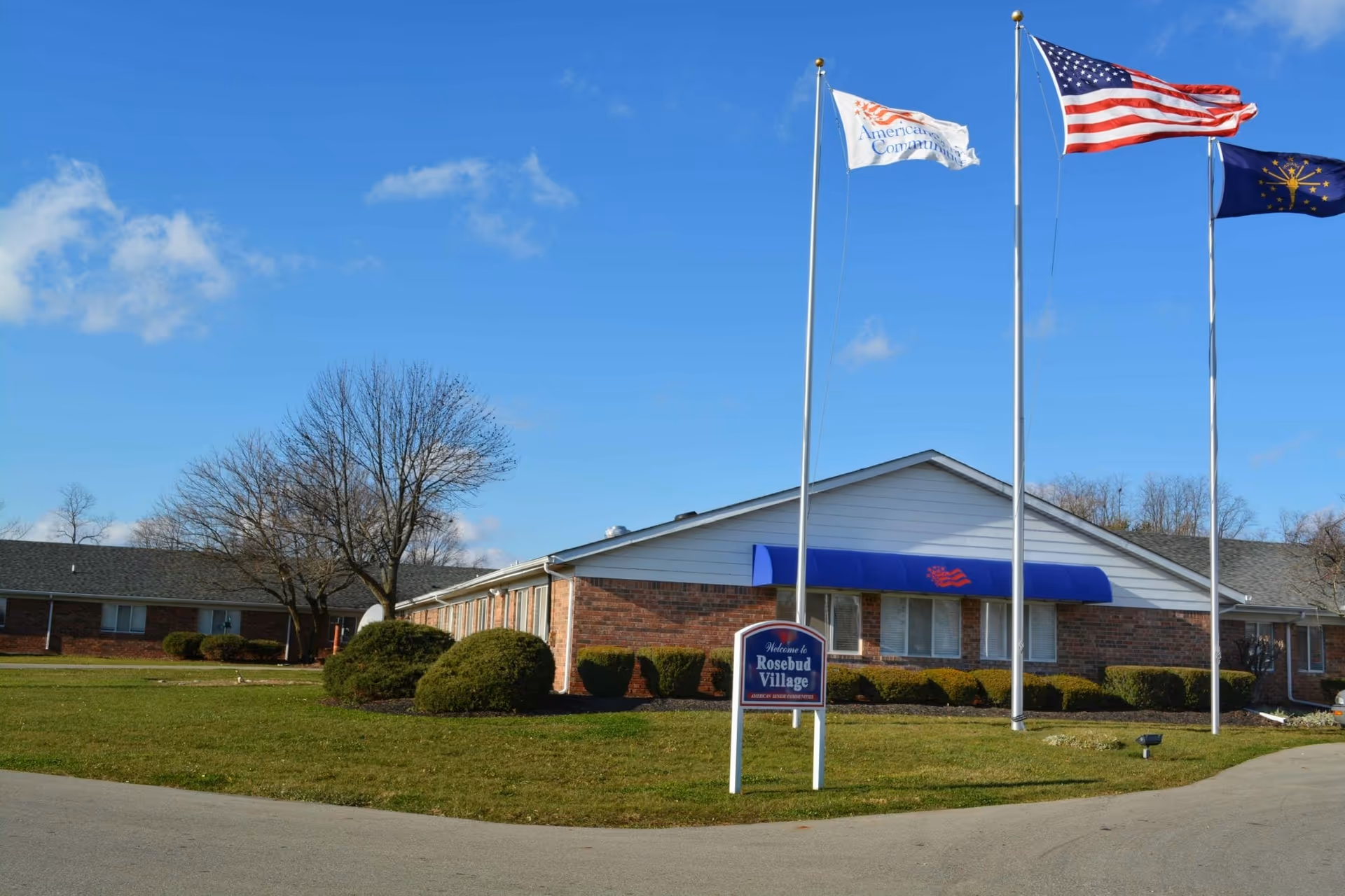 Single-story brick senior living building labeled 'Rosebud Village' with flagpoles and a blue awning on a grassy lawn under a clear sky.