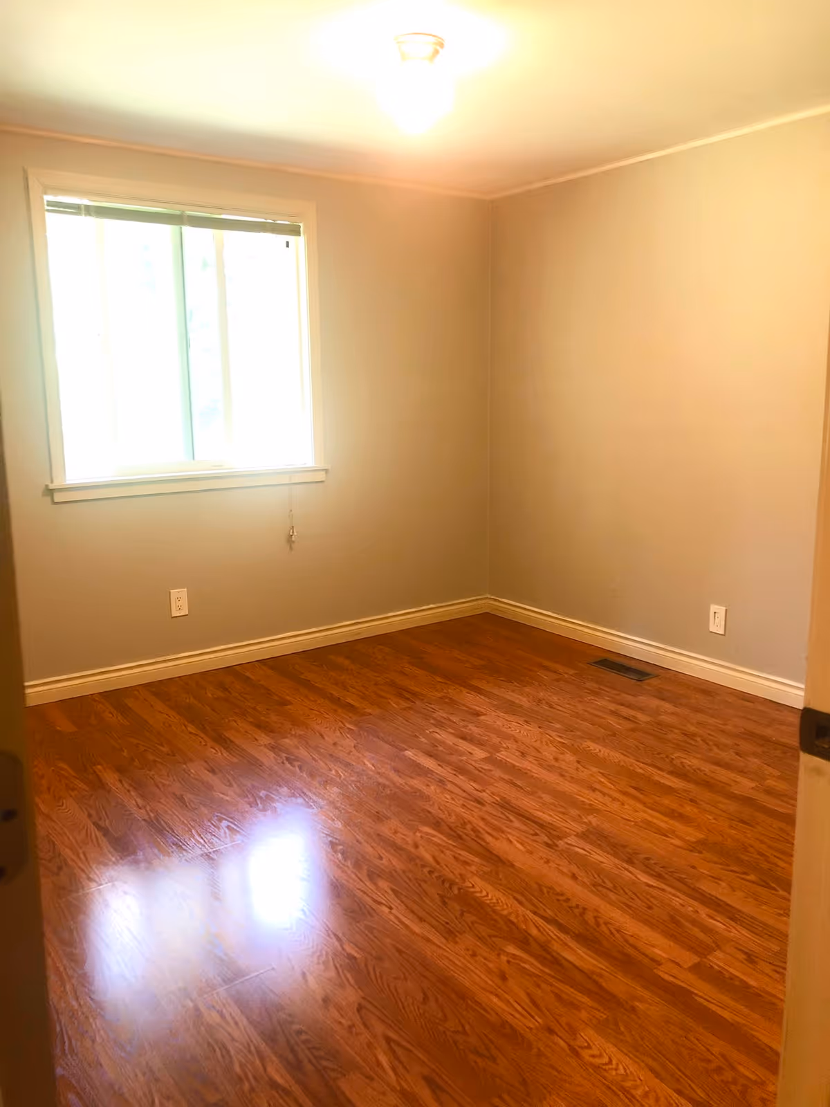 Empty room with wooden flooring, beige walls, a window with blinds, and a ceiling light fixture.