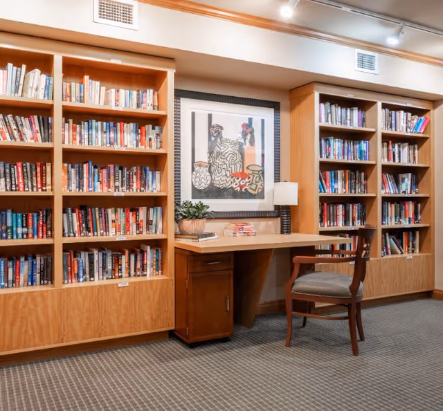 A cozy library or reading area with wooden bookshelves filled with books on either side of a wooden desk. On the desk, there is a small potted plant, a stack of books, and a table lamp. Above the desk hangs a framed abstract artwork. A wooden chair with a cushioned seat is placed in front of the desk. The floor is carpeted with a patterned design.