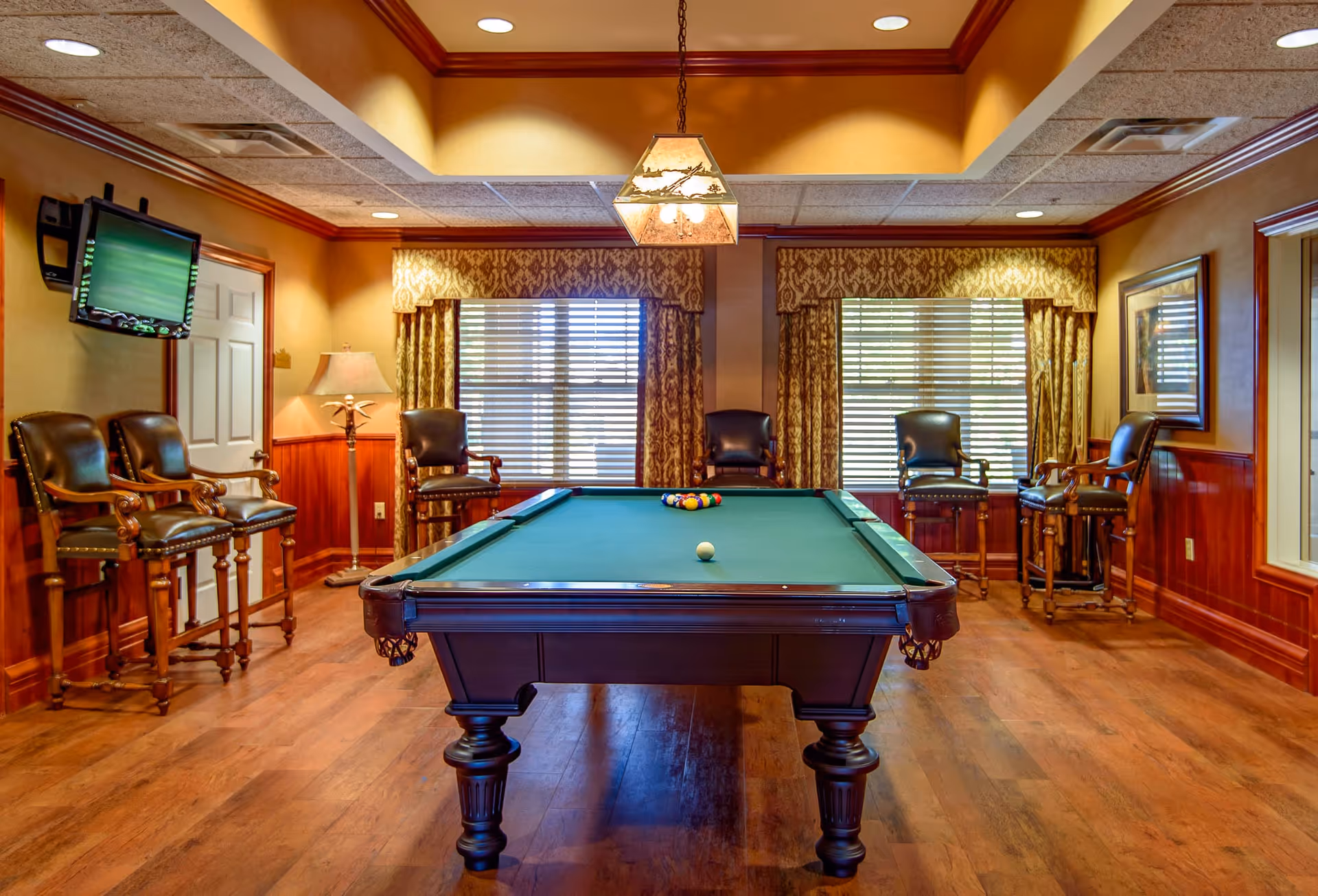 A well-lit game room with a pool table in the center, surrounded by six leather chairs. The room has wooden flooring, wood-paneled walls, two large windows with patterned curtains, a hanging light fixture above the pool table, a floor lamp in the corner, and a wall-mounted TV on the left side.