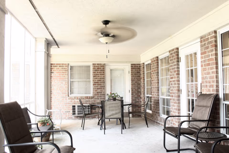 Covered outdoor patio area with brick walls, several cushioned chairs, a round table with four chairs, a ceiling fan, and potted plants.