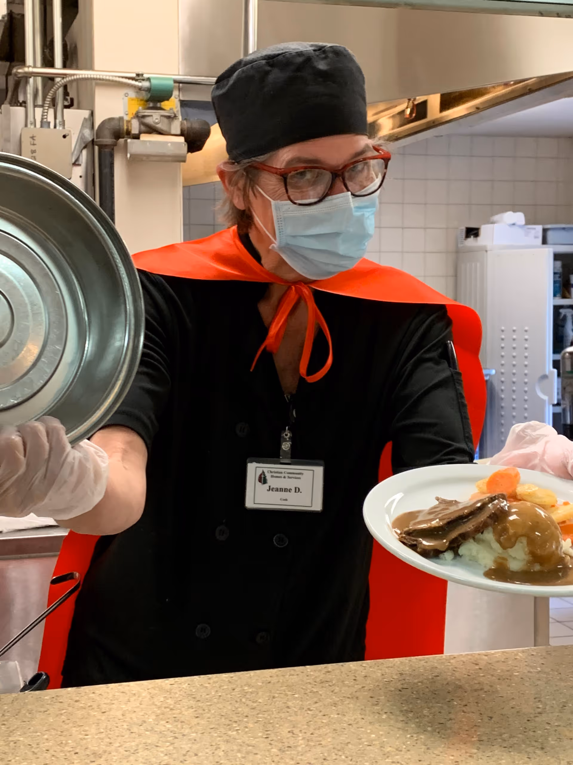 A masked staff member in a black uniform and red cape serves a plate of meat with mashed potatoes and gravy over a cafeteria counter.