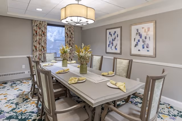 A dining room with a rectangular table set for six people, featuring plates with folded yellow napkins. The table has two vases with floral arrangements as centerpieces. The room has patterned carpet, two abstract paintings on the wall, a window with floral curtains, and a large ceiling light fixture.