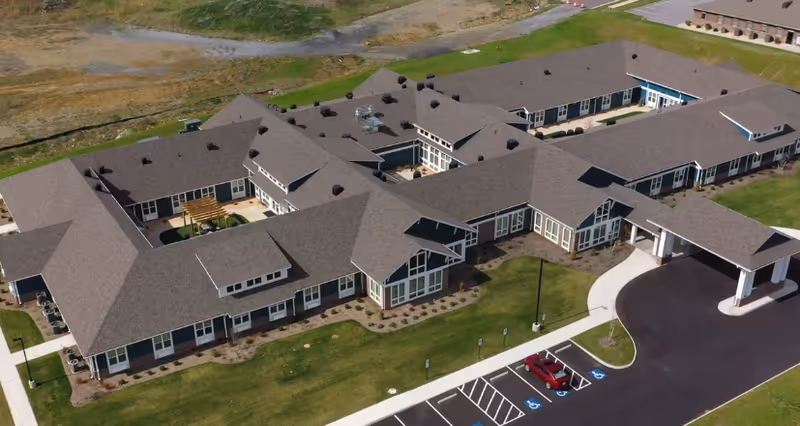 Aerial view of a large single-story senior living facility building with multiple wings and a covered entrance. The building is surrounded by green lawns and a parking lot with several handicap parking spaces and a few cars parked. The surrounding area includes some paved paths and open land.