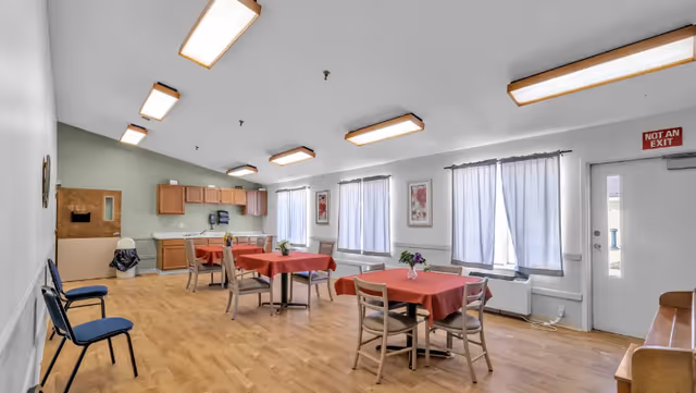 Bright communal dining room with several small tables draped in red tablecloths, chairs, a kitchenette along the back wall, and curtained windows.