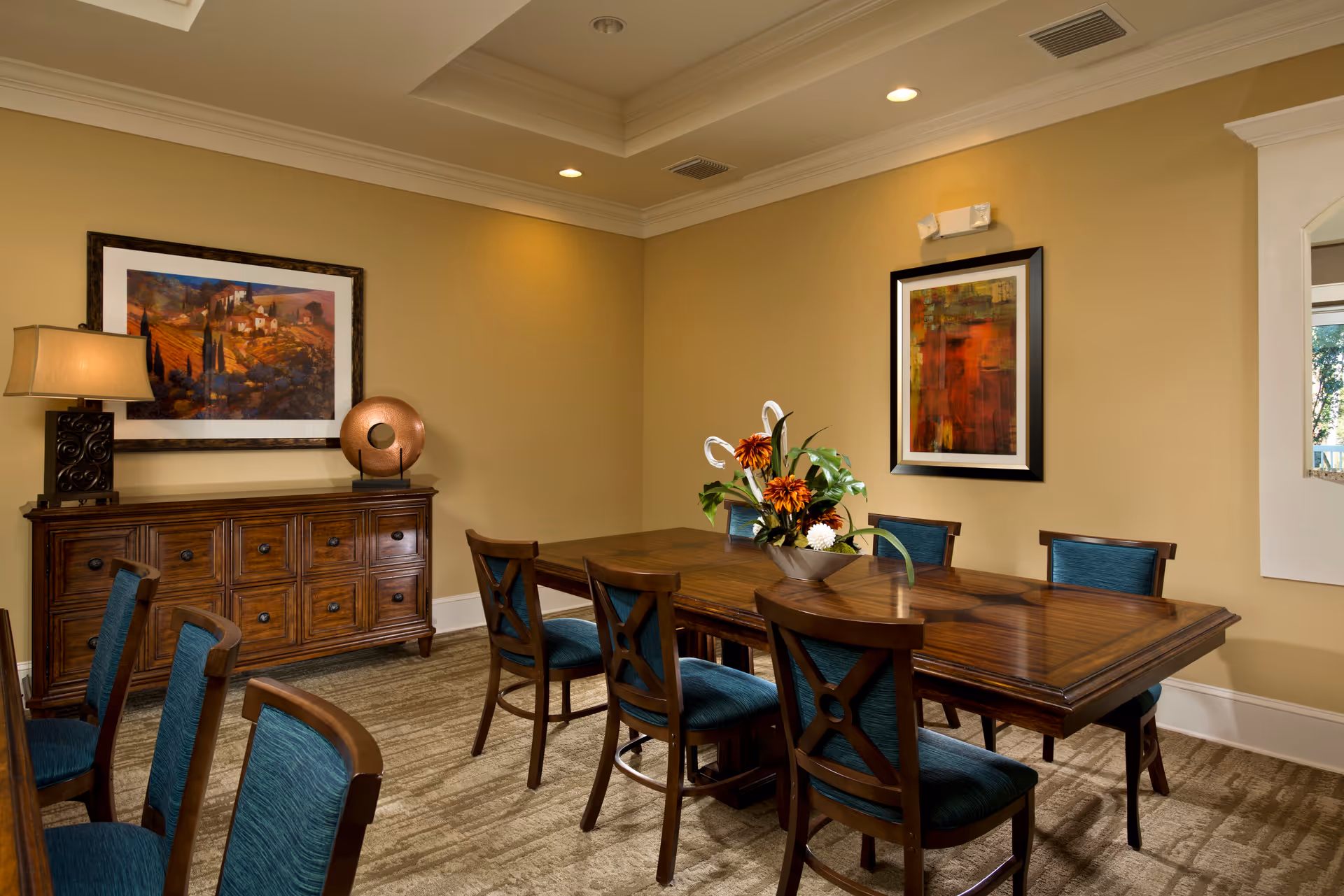 A dining room with a wooden table surrounded by six chairs with blue upholstery. A wooden sideboard with drawers is against the wall, topped with a lamp and decorative item. Two framed artworks hang on the beige walls, and a floral centerpiece decorates the table.