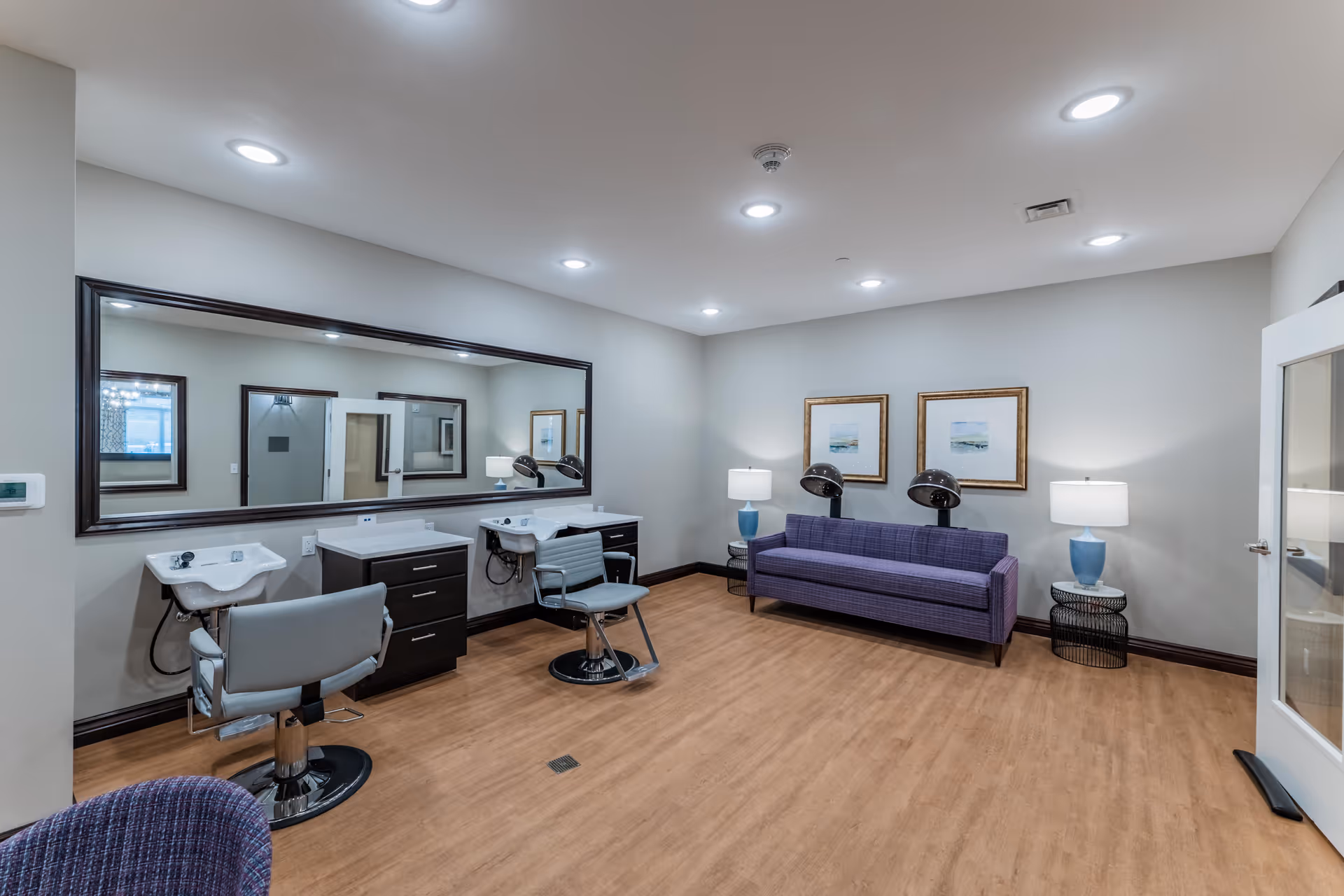 Interior of a senior living facility hair salon area with two salon chairs and sinks in front of a large mirror, a purple couch with two hair dryers behind it, two side tables with blue lamps, and framed artwork on the wall.